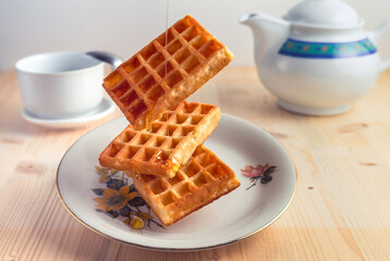 Front view of flying waffles with honey drops on a plate, tea cup with spoon and a tea pot over a wooden table