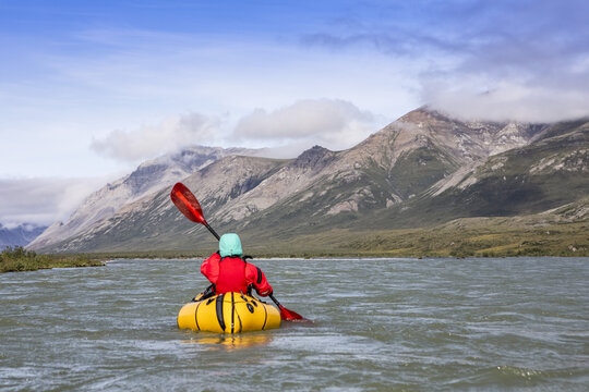 Woman wearing red drysuit and pfd, paddling yellow packraft boat down the Canning river through the Brooks Range, Arctic National Wildlife Refuge; Alaska, United States of America