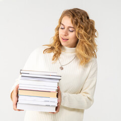 A young attractive blonde woman holds a stack of books in her hands. The girl is dressed in a knitted white sweater. The student rejoices at the passed session and smiles