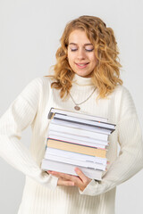 A young attractive blonde woman holds a stack of books in her hands. The girl is dressed in a knitted white sweater. The student rejoices at the passed session and smiles