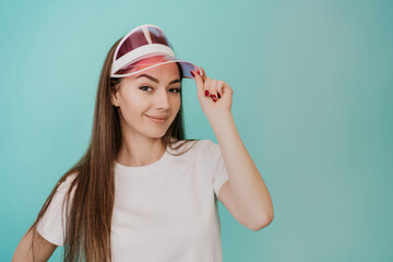 Girl with coloured transparent visor and white t-shirt looks at camera against turquoise studio backdrop. Pretty Italian young sporty fit woman isolated. Mockup, sport, Fitness, healthy lifestyle.