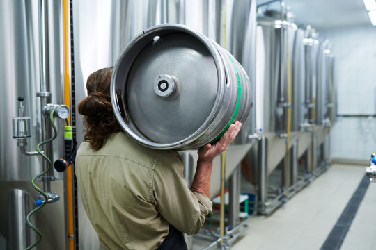 Microbrewery worker carrying beer keg to warehouse, view from back - Powered by Adobe