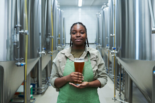 Portrait of positive female worker in uniform showing glass of beer produced in her brewery