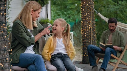 Caucasian funny mother woman with little child girl daughter kid talking speaking laugh mom touch nose of small baby kid talk having fun in camping father reading book read literature on background