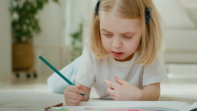 Cute concentrated Caucasian little kid girl draw color pencil lying on floor at home play alone. Talented preschool child daughter kid baby coloring picture at home drawing painting creative paint
