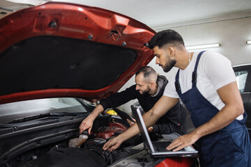 Two young male mechanic using laptop and tablet, recording automobile engine checking collecting detailed information during their work on car workshop. Service maintenance during engine repair.