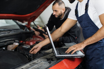 Close up view of hand of young male mechanic using laptop and his colleague with tablet, recording...