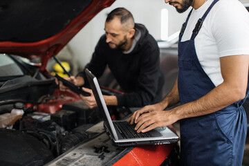 Cropped view of bearded men colleagues car technician mechanics repairing car problem of engine, during system checking detail, using laptop computer for maintenance and fixing in car garage.