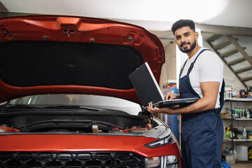 Young bearded man mechanic using laptop for auto diagnostics of sport red car engine at the service...