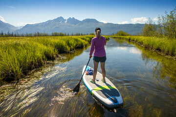 View from behind of a caucasian woman paddle boarding at Rabbit Slough, towards Twin Peaks in the Chugach Mountains, on a sunny, summer day with a blue sky in Wasilla; Alaska, United States of America