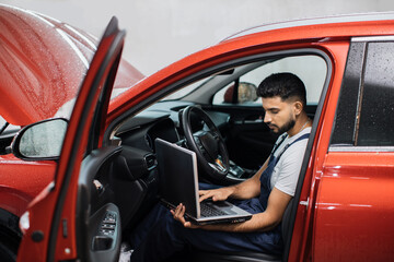 Bearded male mechanic sitting inside car using laptop, recording automobile engine checks collect detailed information during his work on car workshop. Service maintenance during engine repair.