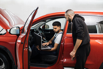 Two young male mechanic using laptop and tablet, recording automobile engine checking collecting detailed information during his work on car workshop. Service maintenance during engine repair.