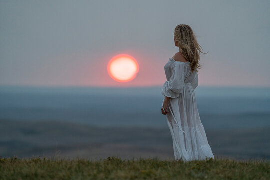 Woman standing on the top of a bluff wearing a white dress while watching the sun setting; Saskatchewan, Canada