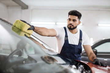 Young bearded man, car wash worker, wearing t-shirt and overalls cleaning the windshield of car...