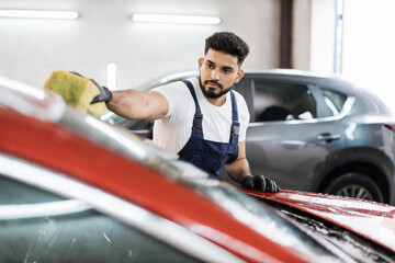 Young bearded man, car wash worker, wearing t-shirt and overalls cleaning the windshield of car...
