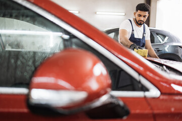 Car wash and clean with shampoo and sponge. Young concentrated bearded man worker, wearing blue...