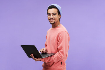 Young successful male freelancer or programmer in pink pullover, beanie hat and eyeglasses holding...