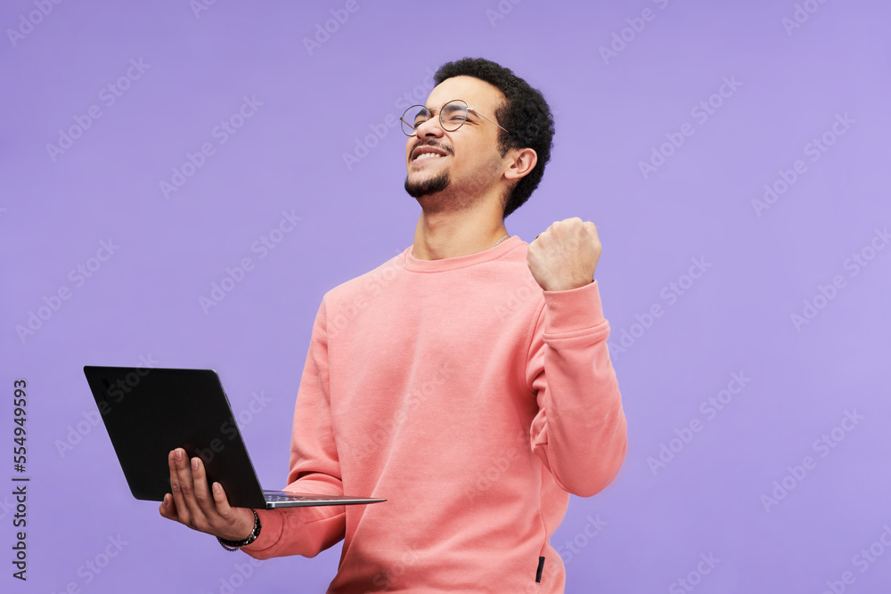Poster ecstatic guy in pink casualwear keeping eyes closed while holding laptop and expressing excitement a