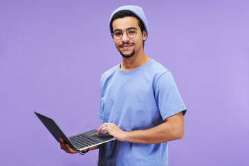 Young successful student or designer in blue t-shirt and beanie hat holding laptop while looking at camera against lavender background - obrazy, fototapety, plakaty