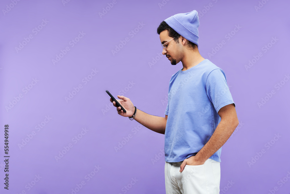 Aufkleber side view of serious guy in blue t-shirt and beanie hat texting in mobile phone or looking through o