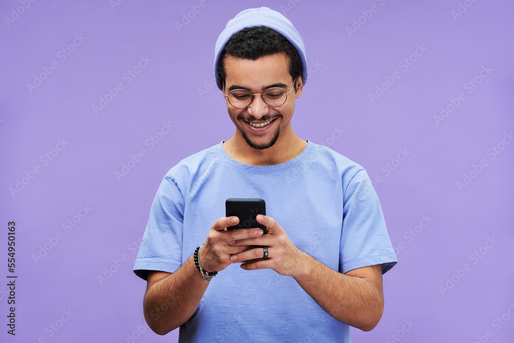Poster young cheerful student in blue t-shirt and beanie hat watching funny video in mobile phone while sta