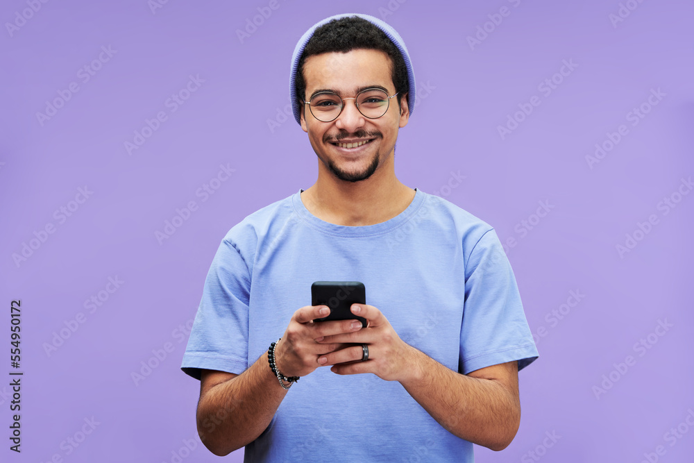 Aufkleber young smiling man in blue casualwear looking at camera while texting in smartphone or scrolling thro