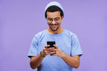 Young cheerful student in blue t-shirt and beanie hat watching funny video in mobile phone while...