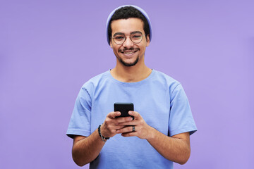 Young smiling man in blue casualwear looking at camera while texting in smartphone or scrolling...