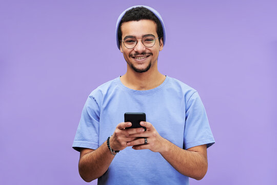 Young smiling man in blue casualwear looking at camera while texting in smartphone or scrolling through photos against violet background