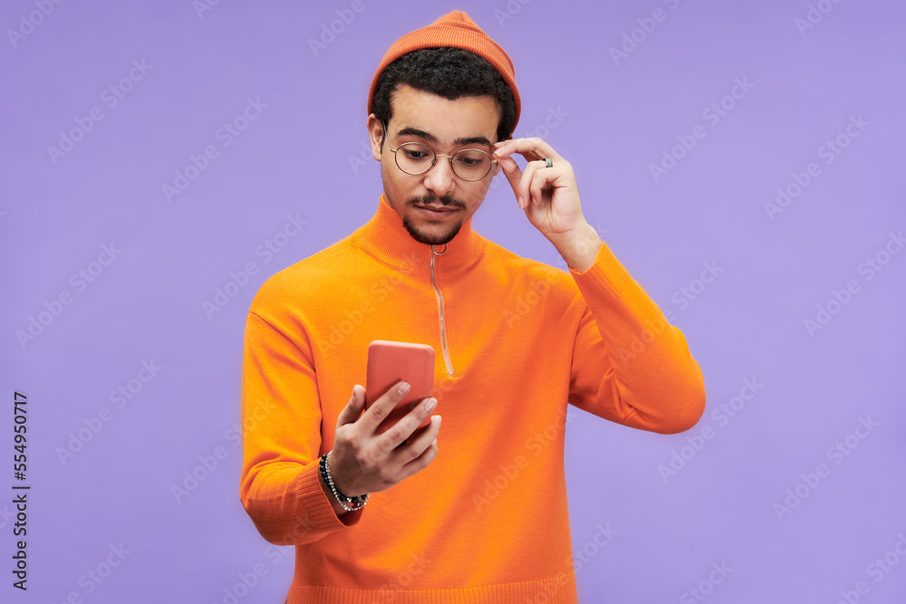 Poster young man expressing misunderstanding while touching eyeglasses and looking at smartphone screen wit