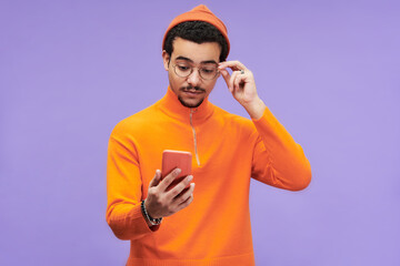Young man expressing misunderstanding while touching eyeglasses and looking at smartphone screen with advert or sale announcement