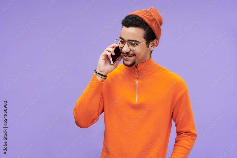 Poster happy guy in eyeglasses, orange pullover and beanie hat talking on mobile phone in front of camera a