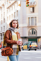 Fototapeta premium vertical portrait of a trendy blonde woman waiting public transportation in the street. She holds a reusable coffee mug
