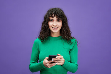 Young woman with dark long wavy hair looking at camera and using mobile phone while standing in isolation on violet background