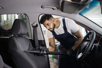 Car wash and detailing service. Bearded male worker in overalls and gloves, making chemical cleaning process with a vacuum cleaner, releasing car seat from dust and sand. - Powered by Adobe