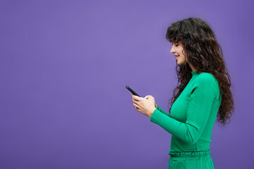 Side view of young brunette woman in green costume using mobile phone while texting or looking...