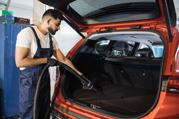 Bearded male worker wearing white t-shirt and blue overalls, vacuuming car interior, trunk with wet vacuum cleaner, professionally extraction method. Wet car vacuum cleaning. © sofiko14