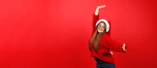 Happy young woman in headphones dancing on a red background in a Christmas sweater and a Santa hat.