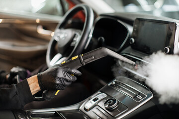 Fototapeta na wymiar Close up of hands of man in black protective rubber gloves cleaning interior of the car with hot steam cleaner. Selective focus on guy hands. Auto cleaning service and detailing concept.