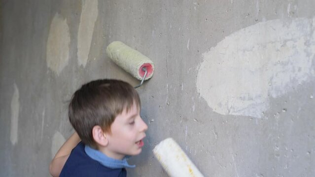 positive caucasian boy 7-8 years old sings and dances near the repaired wall in the apartment holding paint rollers in his hands. selective focus. children participate in the repair of the house