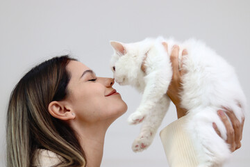 Young happy smiling woman holding cute white Persian cat and lift up her cat, try to kiss her pet, happy beautiful lady playing with fluffy pet, girl hugging her adorable long hair kitty with love