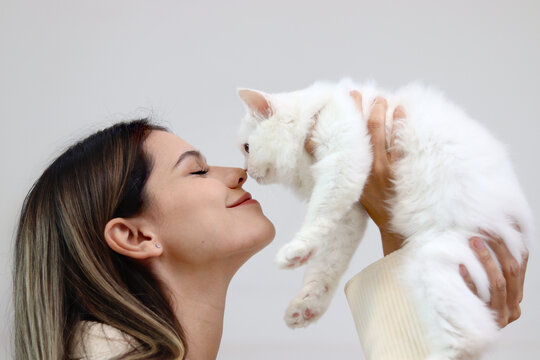 Young happy smiling woman holding cute white Persian cat and lift up her cat, try to kiss her pet, happy beautiful lady playing with fluffy pet, girl hugging her adorable long hair kitty with love
