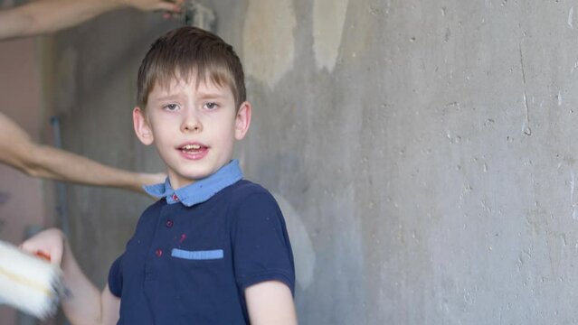 a funny boy of European appearance is dancing with paint rollers looking at the camera, in the background in defocus, his father is using spatulas on the walls, repairs in the house