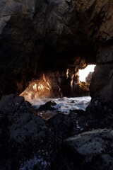 Waves rolling in from the pacific ocean and crashing into rocks at the entrance of a cave in the rays of the sun