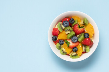 Bowl with fruit salad on blue background. Strawberry, blueberry, kiwi, orange, apple slices. Healthy quick snack concept