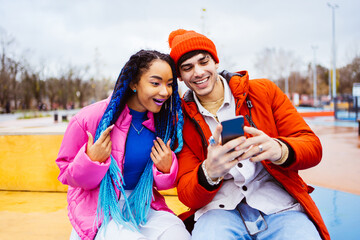 Multiracial young couple of lovers wearing winter jackets dating outdoors in winter - Cool teens using social media app on smartphone - obrazy, fototapety, plakaty