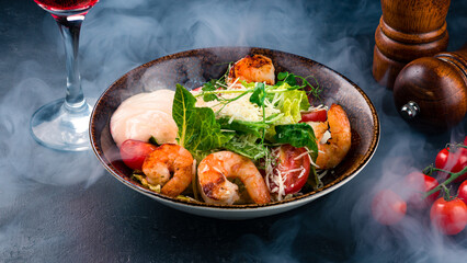 Italian food Caesar salad with shrimps, parmesan cheese, lettuce, tomatoes and sauce on dark table.