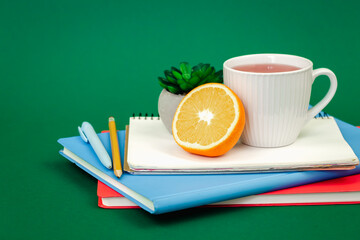 Notebooks, a cup of tea and an orange on a green background, copy space.
