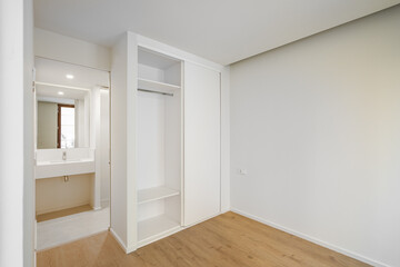 Fototapeta na wymiar Bright spacious bedroom with parquet flooring with a built-in wardrobe for clothes and linen. An open door from the room leading to a bathroom with a washbasin and a small mirror.