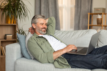 Smiling elderly caucasian man typing on computer, resting, lying on sofa and watching video in living room